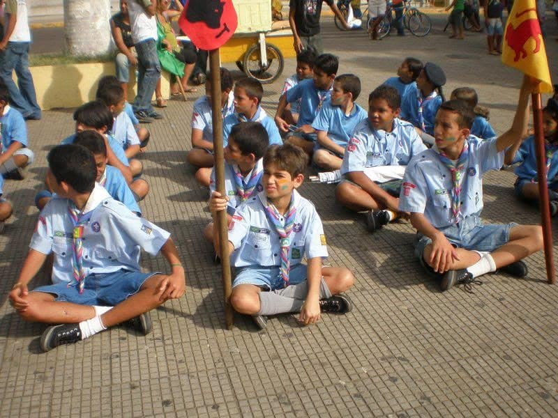 Grupo de escoteiros sentados no chão durante um desfile, com Felipe entre eles